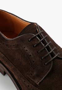 Zapato de vestir de ante marrón con detalles de brogue, puntera redonda y sistema de cordones cerrado. Presenta un forro de cuero suave y una suela texturizada.