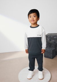 Das Kinderoutfit besteht aus einem grauen und weißen, farblich abgesetzten Sweatshirt mit Rundhalsausschnitt und passenden grauen Jogginghosen, kombiniert mit weißen Turnschuhen.