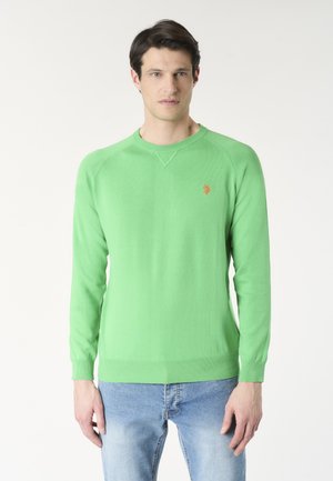 Uomo che indossa un maglione verde brillante a maniche lunghe con un piccolo logo arancione e jeans azzurri, in piedi davanti a uno sfondo bianco semplice.