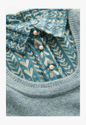 Pullover mit einer weichen blauen Stricktextur; darunter ein gemusterter hellblauer und cremfarbener Kragen mit floralen Motiven und drei Perlenknöpfen.