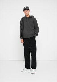 Tânăr îmbrăcat într-un hanorac închis la culoare, pantaloni negri, cu o șapcă neagră, adidași albi și o geantă de umăr neagră, stând în fața unui fundal alb.