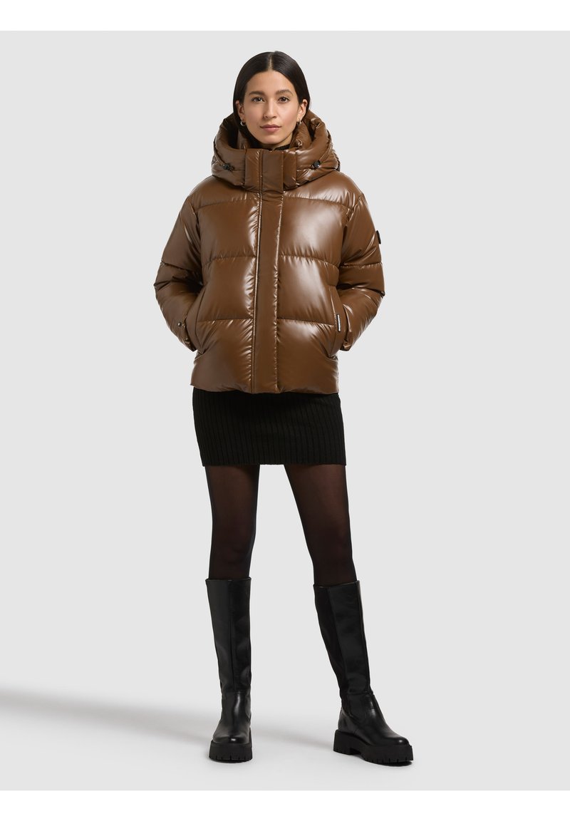 Giacca marrone imbottita con collo alto e cappuccio, caratterizzata da una finitura lucida e cuciture visibili. Abbinata a una gonna nera a costine e stivali al ginocchio.