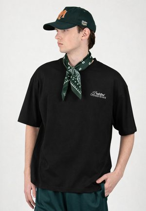 Jonge man die een zwart T-shirt met witte tekst draagt, een donkergroene bandana om zijn nek geknoopt, een zwarte pet met een oranje logo en groene shorts.