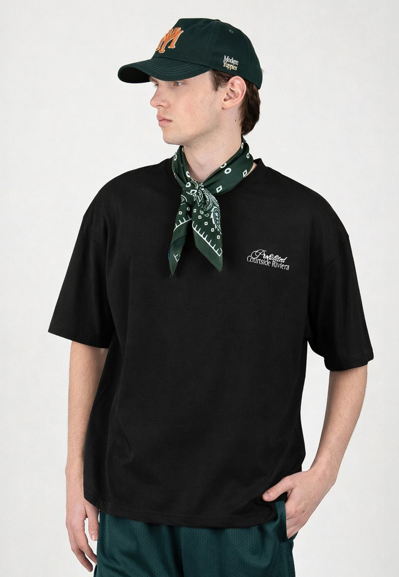 Jeune homme portant un t-shirt noir avec un texte blanc, un bandana vert foncé noué autour du cou, une casquette noire avec un logo orange et un short vert.