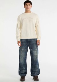 Cream-colored sweater with ribbed cuffs and crew neckline, paired with baggy blue denim cargo pants featuring multiple pockets and textured fabric.