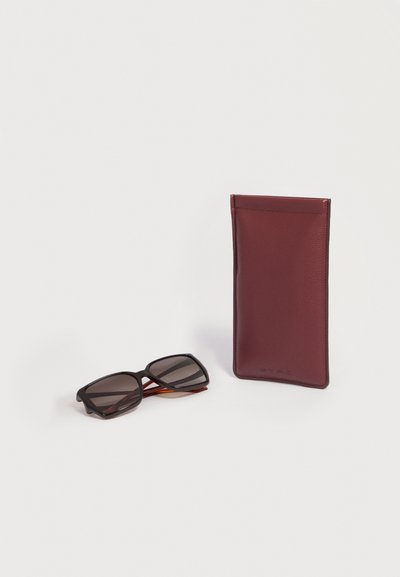 Lunettes de soleil écaille avec verres foncés à côté d'un étui à lunettes vertical en cuir grenat sur une surface blanche.