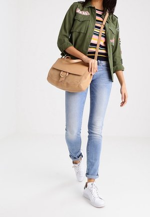 Osoba spacerująca, ubrana w zieloną kurtkę, pasiastą koszulę, jasnoniebieskie dżinsy, białe tenisówki i nosząca beżową torebkę na ramię.