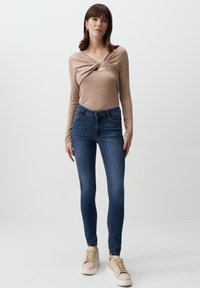 Maglietta beige a maniche lunghe con design a torsione, abbinata a jeans fitted blu scuro e sneaker beige. Abbigliamento semplice e casual con accenti minimi.