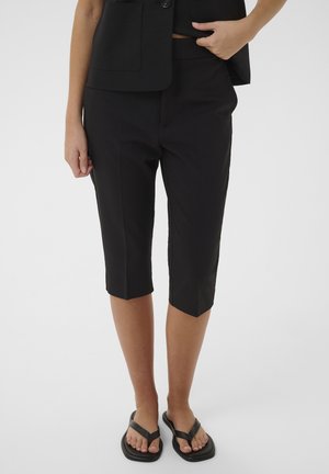 Osoba oblečená v čiernych skrátených na mieru urobených nohaviciach, čiernej veste a čiernych sandáloch s hranatou špičkou, stojaca pred jednoduchým svetlým pozadím.