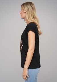 Svart bomull T-skjorte med rullede ermer og blomsterprint i rosa og oransje. Modellen har på seg lyseblå jeans. Profilbilde fra siden.