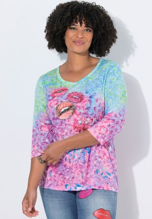 Woman with curly hair wearing a colorful shirt with lip prints and jeans with lip patches, standing against a white wall.