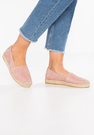Persona que lleva zapatos de esparto rosa claro y vaqueros azules con el dobladillo deshilachado, de pie sobre un suelo blanco con un pie ligeramente levantado.