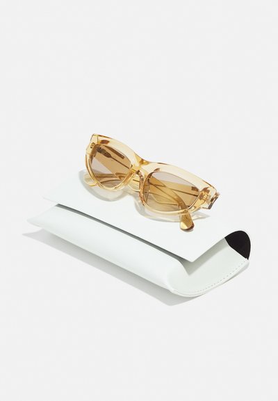 Cat-eye sunglasses with transparent golden frames and brown-tinted lenses resting on a white leather case with a black accent.