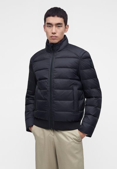 Un homme porte une doudoune matelassée noire avec un col montant zippé, associée à un pantalon beige, les mains dans les poches, sur un fond uni.