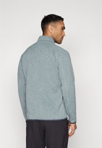 Casaco de polar azul claro com gola alta, tecido texturizado e remate contrastante na bainha. Apresenta um corte relaxado e mangas longas.