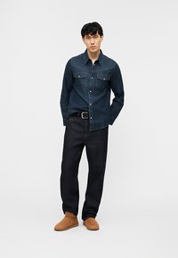 Junger Mann steht und trägt ein dunkles Jeanshemd, dunkle Jeans mit schwarzem Gürtel und hellbraune Wildleder-Stiefeletten vor weißem Hintergrund.