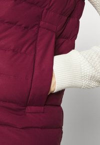 Burgundský prošívaný vest s boční kapsou, který má jemný texturovaný krémový svetr vyčuhující zpod.