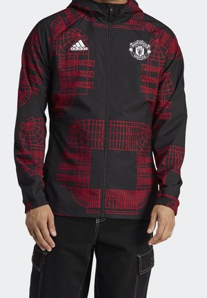 Veste noire zippée avec un motif géométrique rouge de stade, logo Adidas sur la poitrine droite et écusson de Manchester United sur la poitrine gauche.