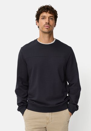 Marineblå crewneck-sweatshirt med lange ærmer, tekstureret stof og en vandret søm over brystet. Kombineret med beige bukser.