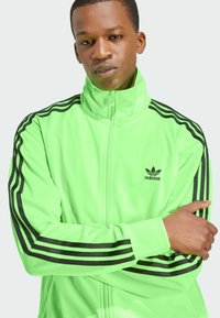 Neon groene Adidas trainingsjack met een hoge kraag, zwarte strepen op de schouders en een klein logo aan de bovenste linkerkant.