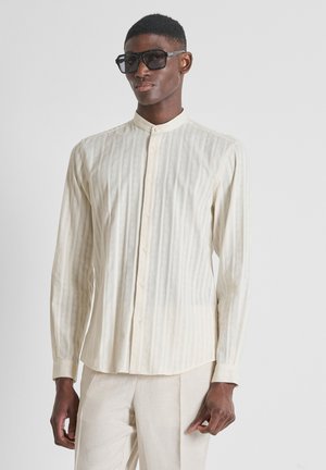 Man draagt een textuurrijk crème button-up shirt met mandarijnkraag, bijpassende lichte broek en oversized zwarte zonnebril, staand tegen een grijze achtergrond.