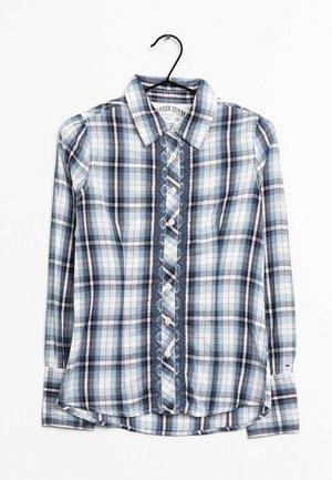 Camisa de cuadros azul y blanca de manga larga con cierre de botones y detalle de encaje, colgada en una percha negra sobre fondo blanco.