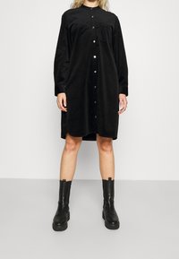 Czarna sukienka koszulowa z korduroy z długimi rękawami, zapięciem na guziki z przodu i kieszenią na piersi. Noszona z czarnymi, wysokimi botkami na platformie.
