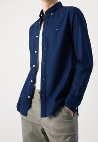 Chemise bleu marine à boutons en coton, dotée d'un col classique, de manches longues et d'un petit logo sur la poitrine. Associée à un pantalon clair.