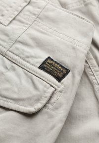 Tissu beige avec couture et poche cousues, portant une étiquette de marque Superdry noire et dorée cousue au-dessus du rabat de la poche.
