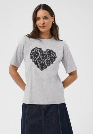 Femme aux longs cheveux portant un t-shirt gris clair avec un grand cœur en dentelle noire et une jupe en jean foncé, debout devant un fond uni.