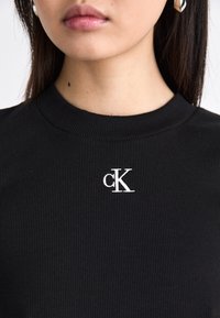 T-shirt noir à côtes avec un col rond, orné d'un logo "cK" brodé en blanc sur la poitrine. Texture lisse, design ajusté.
