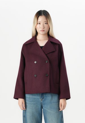 Jeune femme avec des cheveux longs dégradés porte une veste cintrée violette foncée à double boutonnage et un jean bleu devant un fond blanc uni.