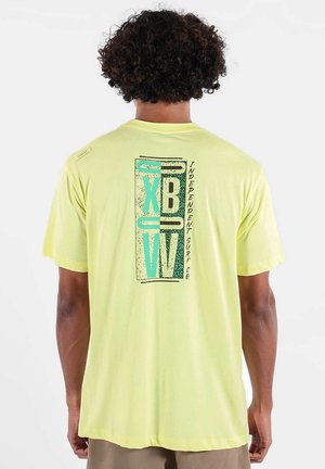 Homme aux cheveux bouclés portant un T-shirt jaune pâle avec un graphisme turquoise et noir "XBW Independent Surf Co." au dos.