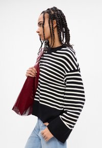 Suéter de malha às riscas pretas e brancas com um corte solto e manga sino, combinado com uma mala de tote burdeos brilhante. Jeans azul claro completam o visual.