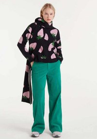 Sudadera negra con estampados de fresas rosas y hojas verdes, combinada con pantalones anchos de pana verde azulado. Los zapatos son blancos y grises.