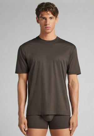 Jonge man met kort krullend haar, draagt een donkerbruine aansluitende T-shirt en bijpassende boxershorts tegen een effen grijze achtergrond.