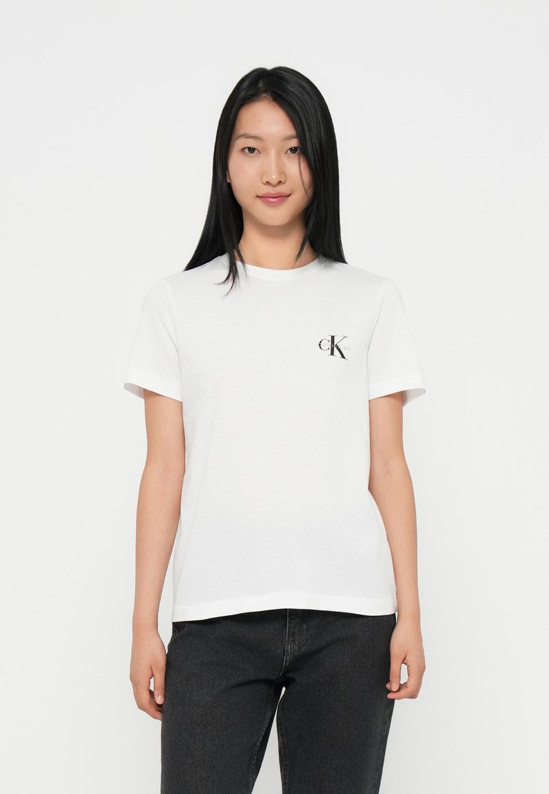 Camiseta de algodón blanca con cuello redondo, mangas cortas y un pequeño logo negro "CK" en la parte superior izquierda. Combinada con jeans oscuros.