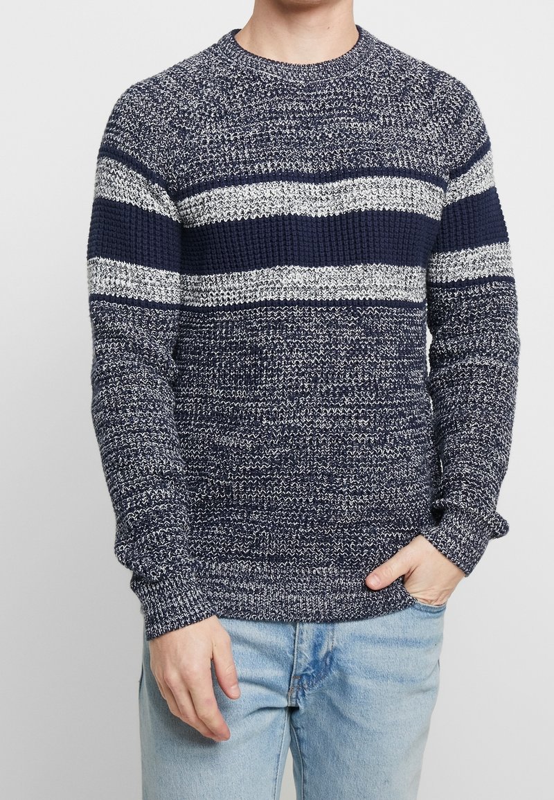 Hombre con un suéter de punto a rayas en navy y blanco y jeans azul claro, de pie con una mano en el bolsillo frente a un fondo liso.