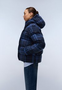 Pufjakke i mørkeblå plaidmønster med hætte, lynlåslukning og sidelommer. Bæres over en hvid skjorte og blå jeans.