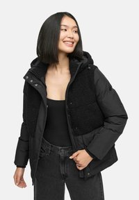 Black puffer jacket with a fluffy black fleece panel on the front. Features a hood, zipper closure, and snap buttons. Worn over a black top.