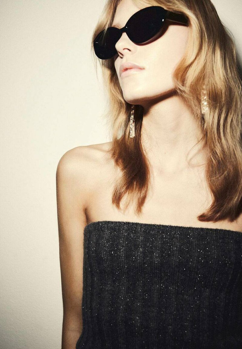 Occhiali da sole neri con montatura spessa, indossati con un top a coste grigio scuro senza spalline. Orecchini pendenti notevoli con un design geometrico.