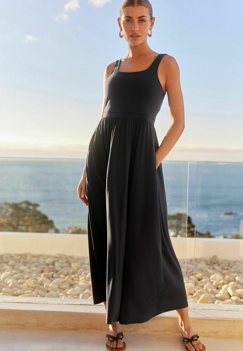 Mörkblå ärmlös maxiklänning med en figurnära bodice, rynkad midja och flödande kjol. Modellen står med händerna i fickorna och har på sig bruna sandaler.