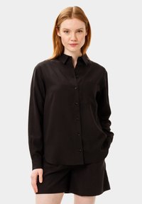 Camicia nera a maniche lunghe con colletto a punta, taschino anteriore e una texture liscia, abbinata a pantaloni corti neri.