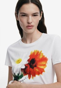 Hvid bomulds t-shirt med et farverigt blomstermotiv, der inkluderer en gul tusindfryd og en orange solsikke, suppleret med en signaturdetalje.