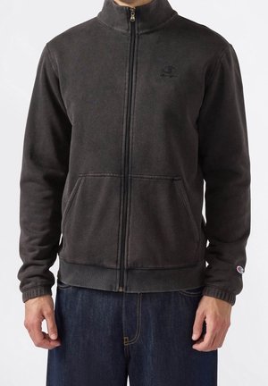 Mand iført en mørkegrå fleecejakke med lynlås og lommer og jeans, stående mod en ensfarvet hvid baggrund.