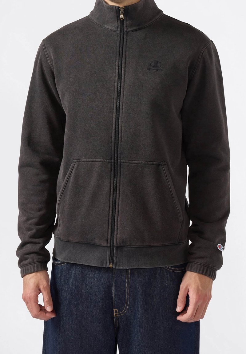 Homme portant une veste polaire zippée gris foncé avec des poches et un jean, debout devant un fond blanc uni.