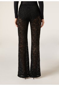 Femme portant un pantalon en dentelle noir taille haute avec un motif floral et des jambes évasées, vue de dos sur un fond uni.