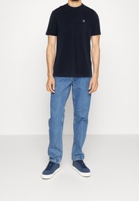 Muž v jednoduchém tmavě modrém tričku, světle modrých džínách a tmavě modrých teniskách stojící na bílém pozadí.