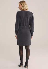 Žena s krátkymi blond vlasmi, oblečená v dlhom tmavošedom textúrovanom šate, čiernych pančuchách a čiernych topánkach na opätku, stojí s chrbtom k nám.