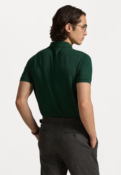 Camisa polo verde escura vista de costas, com mangas curtas e uma textura suave, combinada com calças cinzentas com bolsos traseiros.